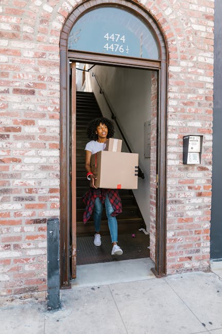 Two young women sitting at the back of an open moving van during a home relocation process, surrounded by packed cardboard boxes and plastic-wrapped furniture. The woman on the left has curly hair and